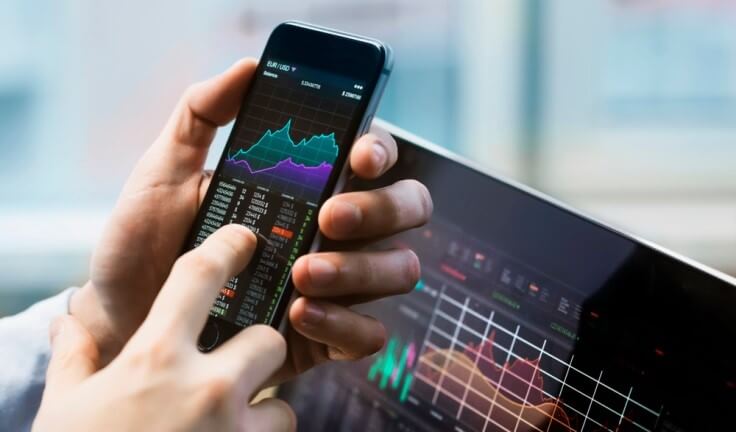Finance Brokerage-Share Market: a hand holding a phone while checking graphs on stock markets.